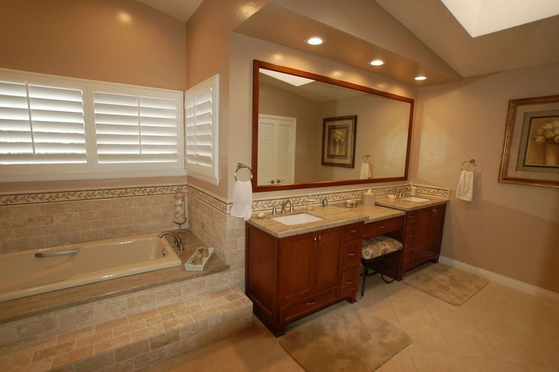 Bathroom with tub, vanity, and large mirror. Beige walls, wood cabinets, and white shutters.