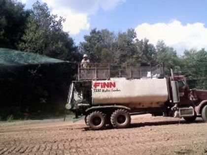 Large tanker truck spraying water on a dirt road beside trees under a blue sky