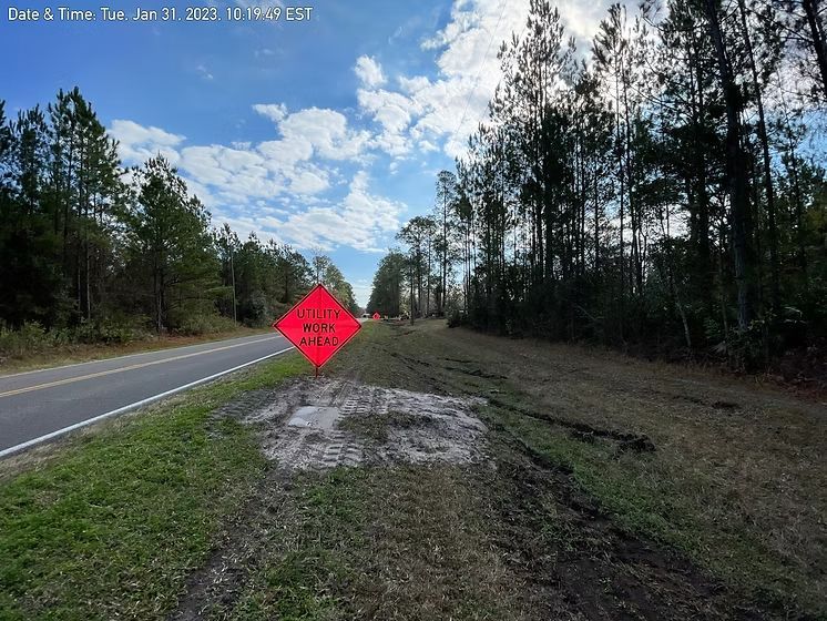 Rural road with a bright red warning sign beside a dirt shoulder and pine trees under a cloudy sky