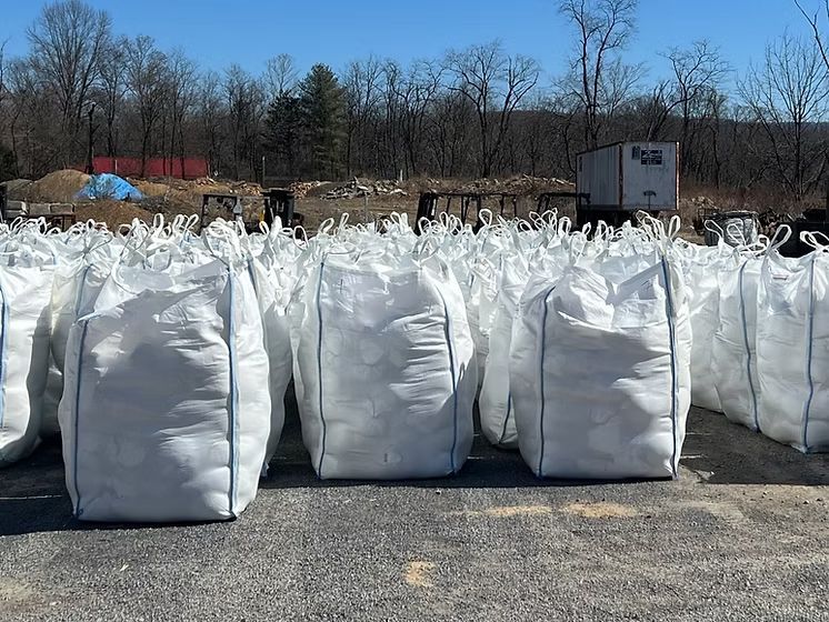 Rows of large white bulk bags stacked outdoors on asphalt near leafless trees under a blue sky