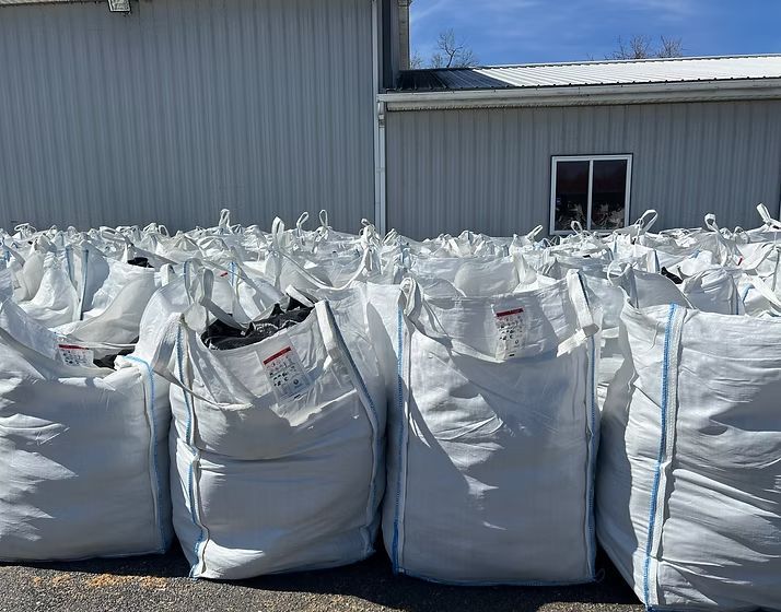 Rows of large white bulk bags piled outdoors beside a gray industrial building under clear sky