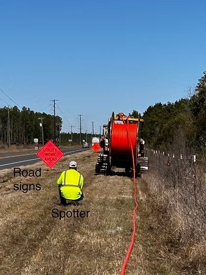 Roadwork scene with a hi-vis spotter, orange cable reel, and warning sign on a rural roadside.