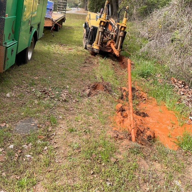 Backhoe digging a muddy trench beside a green utility truck along a grassy roadside