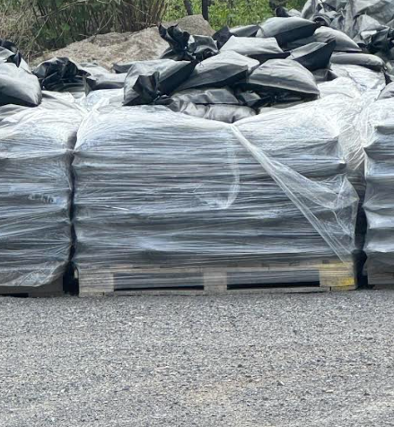 Wrapped pallets of black bags stacked in an outdoor gravel yard with trees behind them