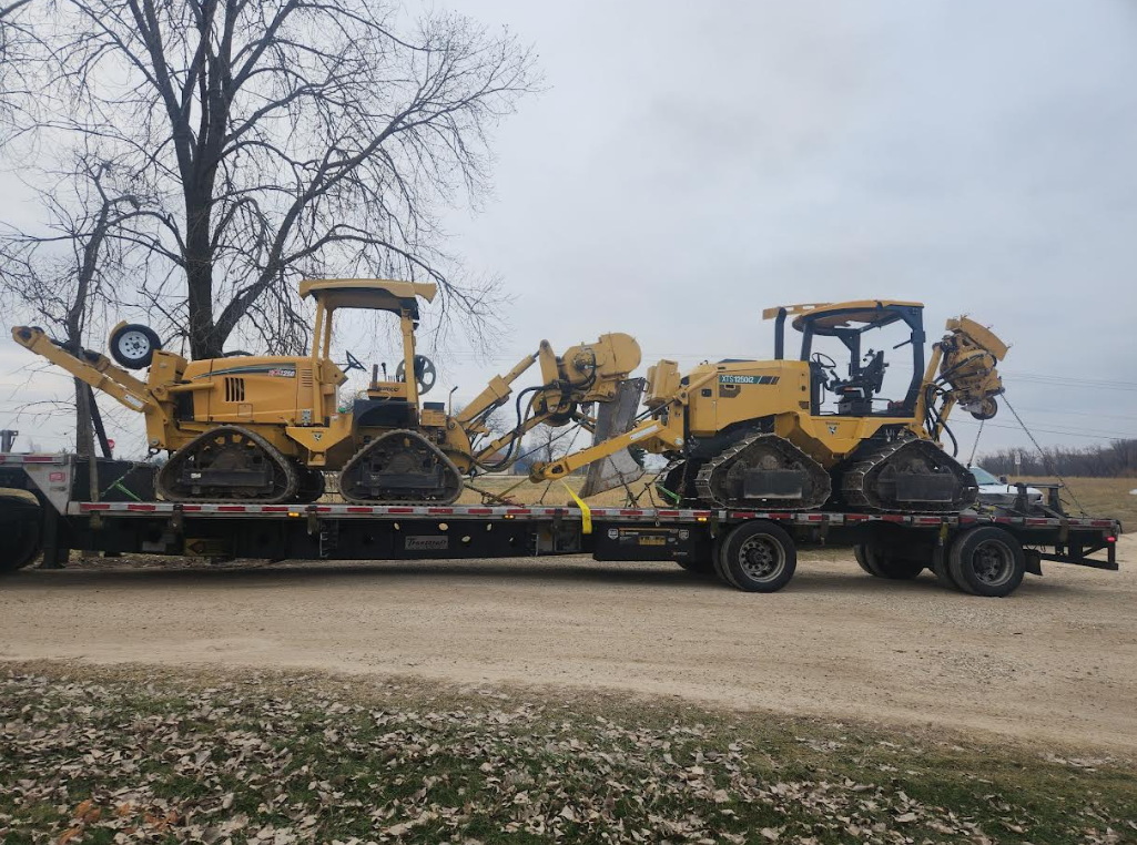 Two yellow construction machines secured on a black trailer beside a leafless tree