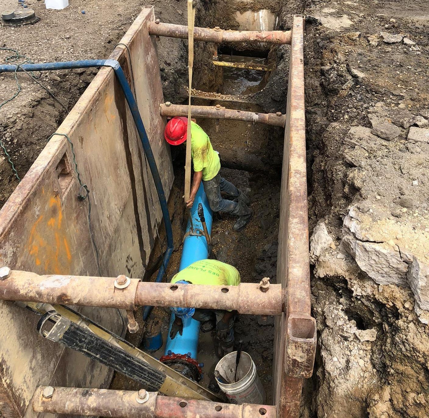 Workers in hard hats and vests repairing blue pipe in a trench, using safety supports.