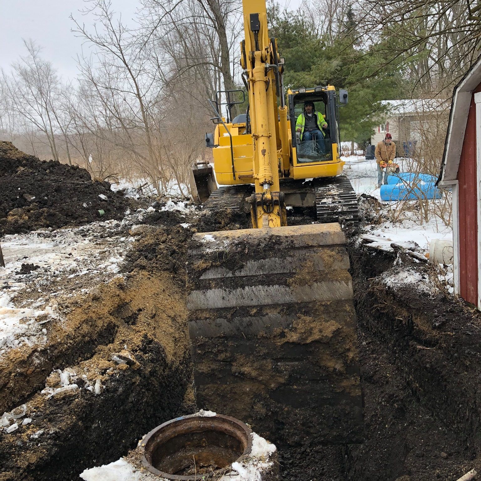 Yellow excavator digging near a metal access hatch in snow. A man operates the machinery.