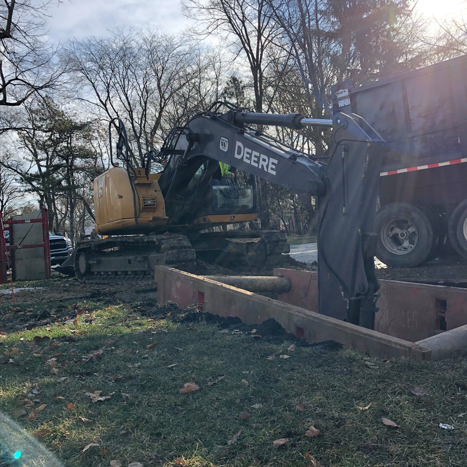 A John Deere excavator digging near a dump truck; brown dirt, bare trees, and a cloudy sky are in the background.