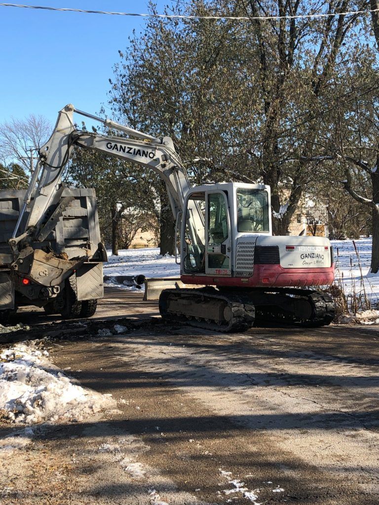Excavator loading a dark truck on a snow-covered road. Blue sky. Bare trees in the background.