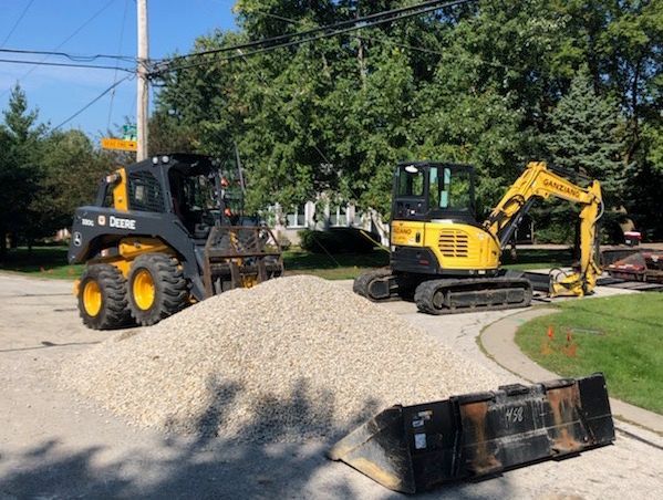 A John Deere skid steer and a yellow excavator near a pile of gravel, likely for road work.