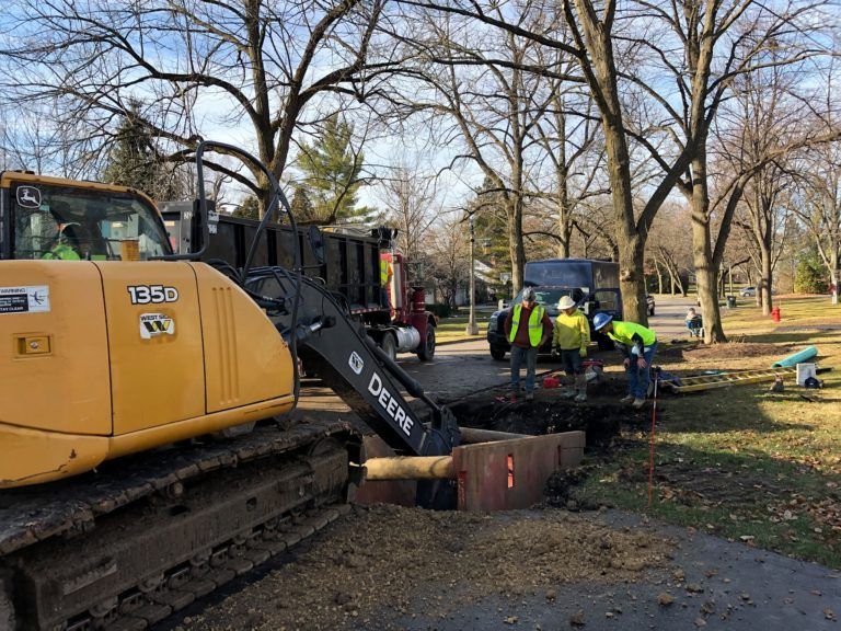 Yellow excavator and crew working on road, digging a trench with a truck in the background.