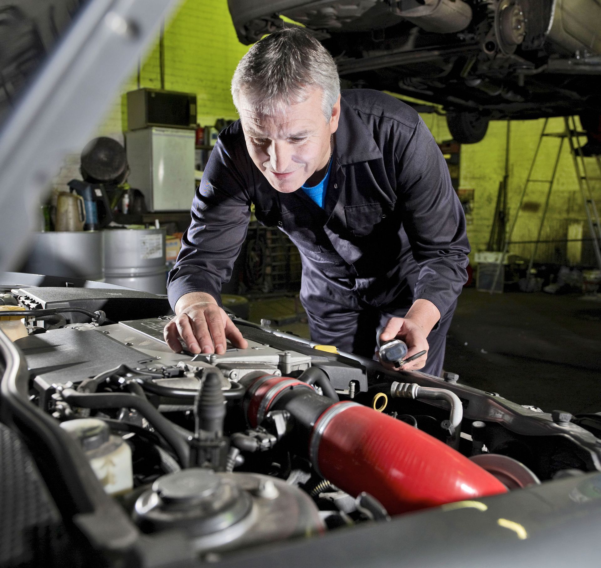 Mechanic inspecting a car engine in a garage, with a red intake hose visible.