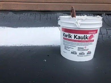 White bucket of Kwik Kaulk with a brush on a black roof surface.