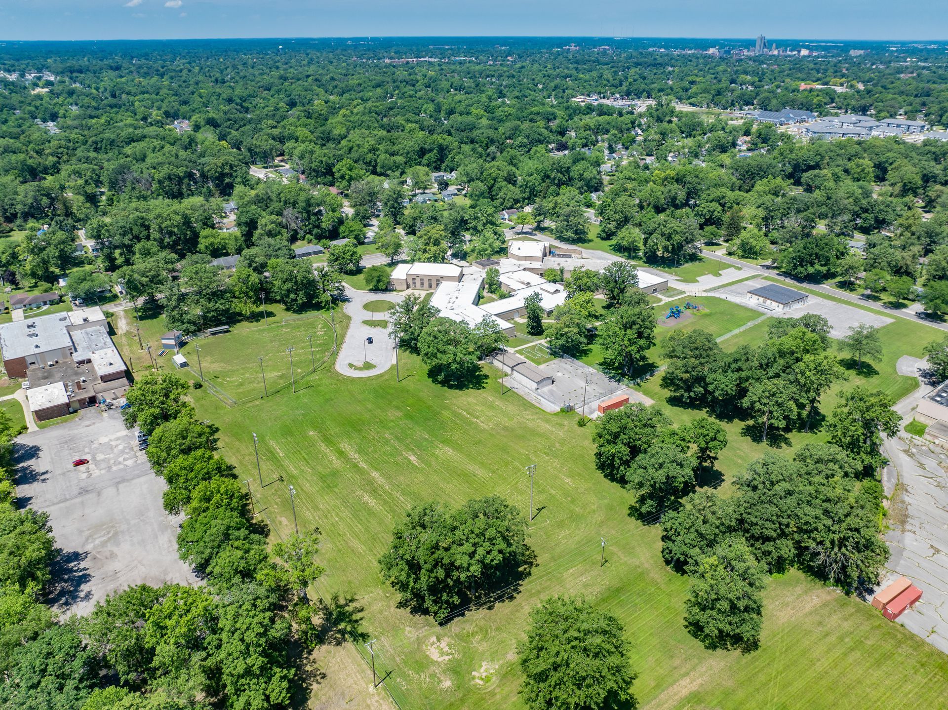 Aerial view of a grassy field with scattered trees and buildings amid a dense green neighborhood.