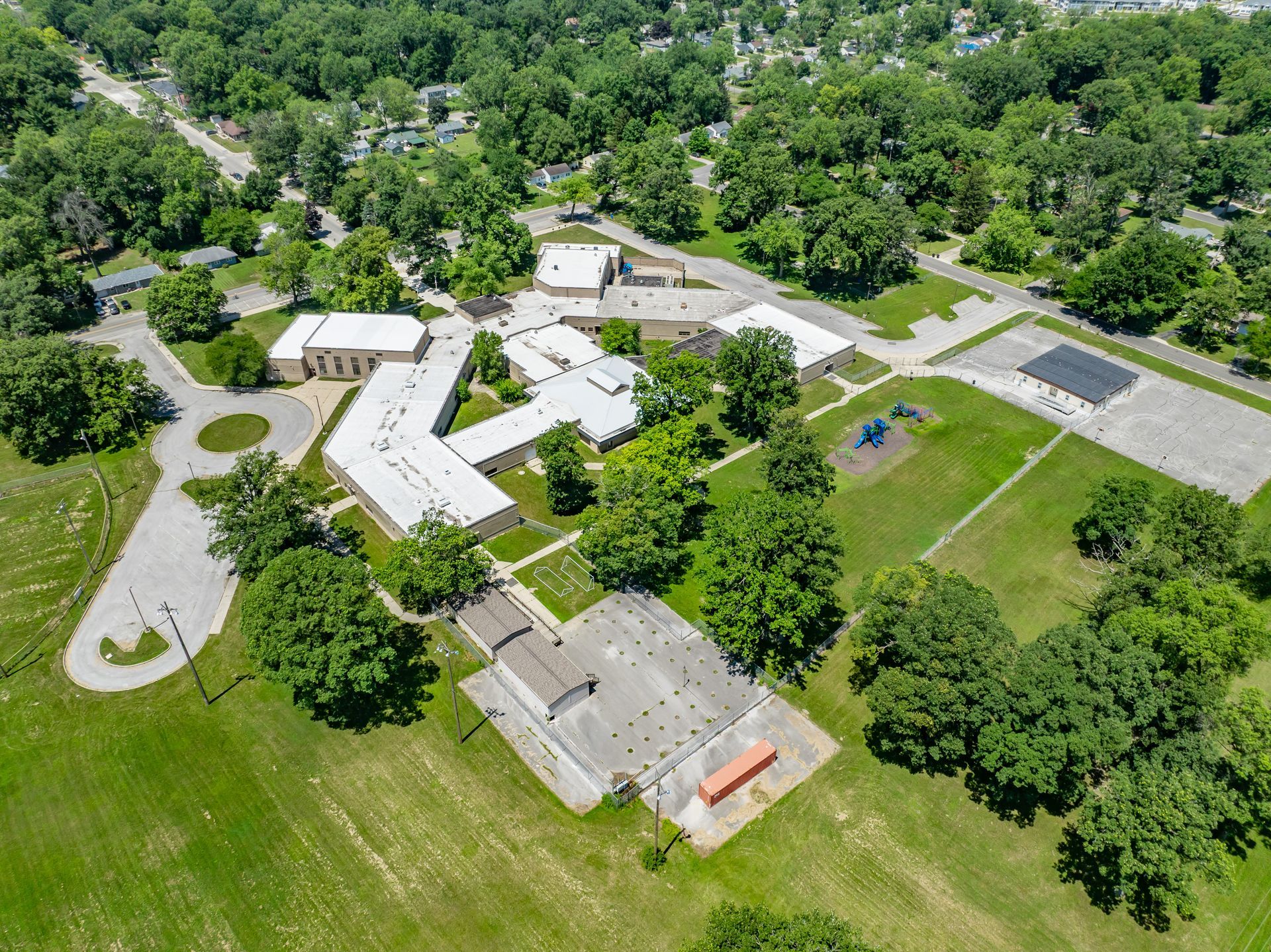Aerial view of a wooded campus with low buildings, roads, parking lots, and open grassy areas.
