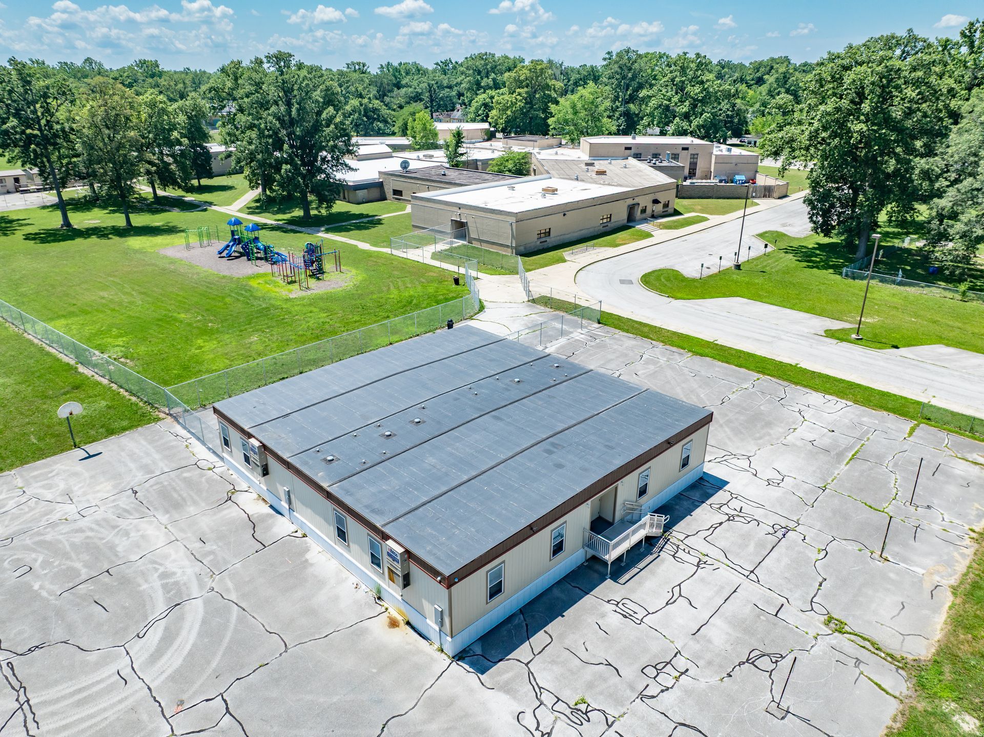 Aerial view of a school campus with a gray-roofed building, parking lot, and playground surrounded by trees