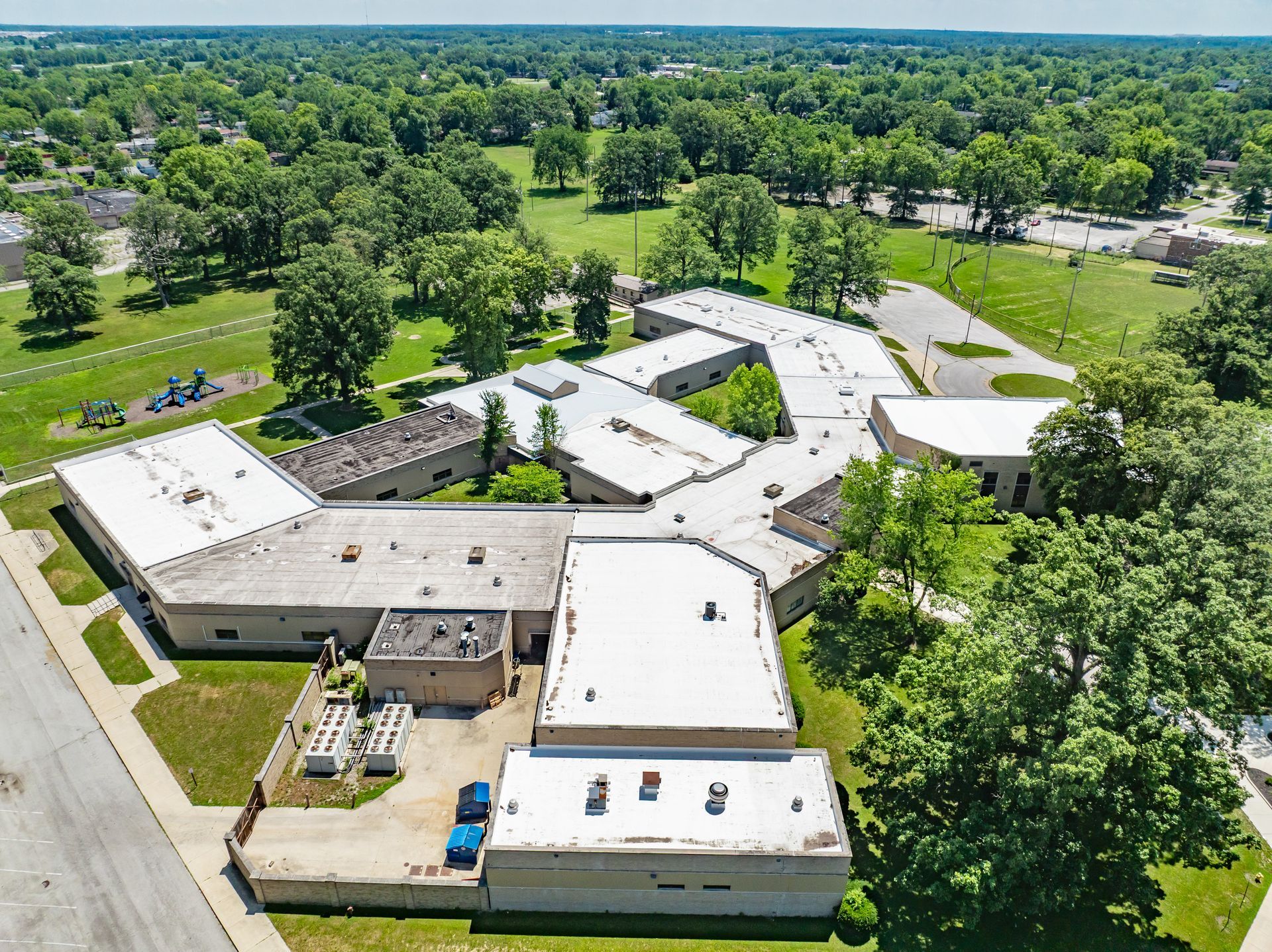 Aerial view of a large, single-story school or community building with a flat, white roof, surrounded by green lawns and trees.