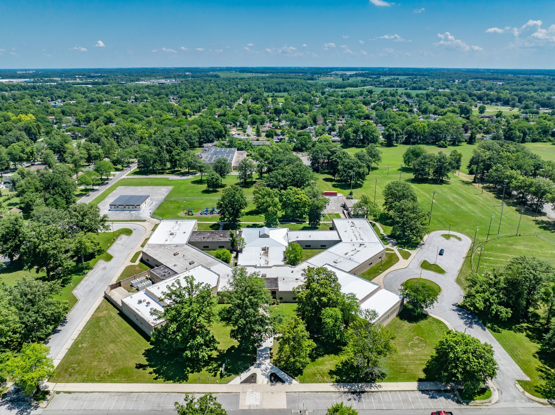 Aerial view of a white-roofed campus complex surrounded by green trees and fields under a blue sky