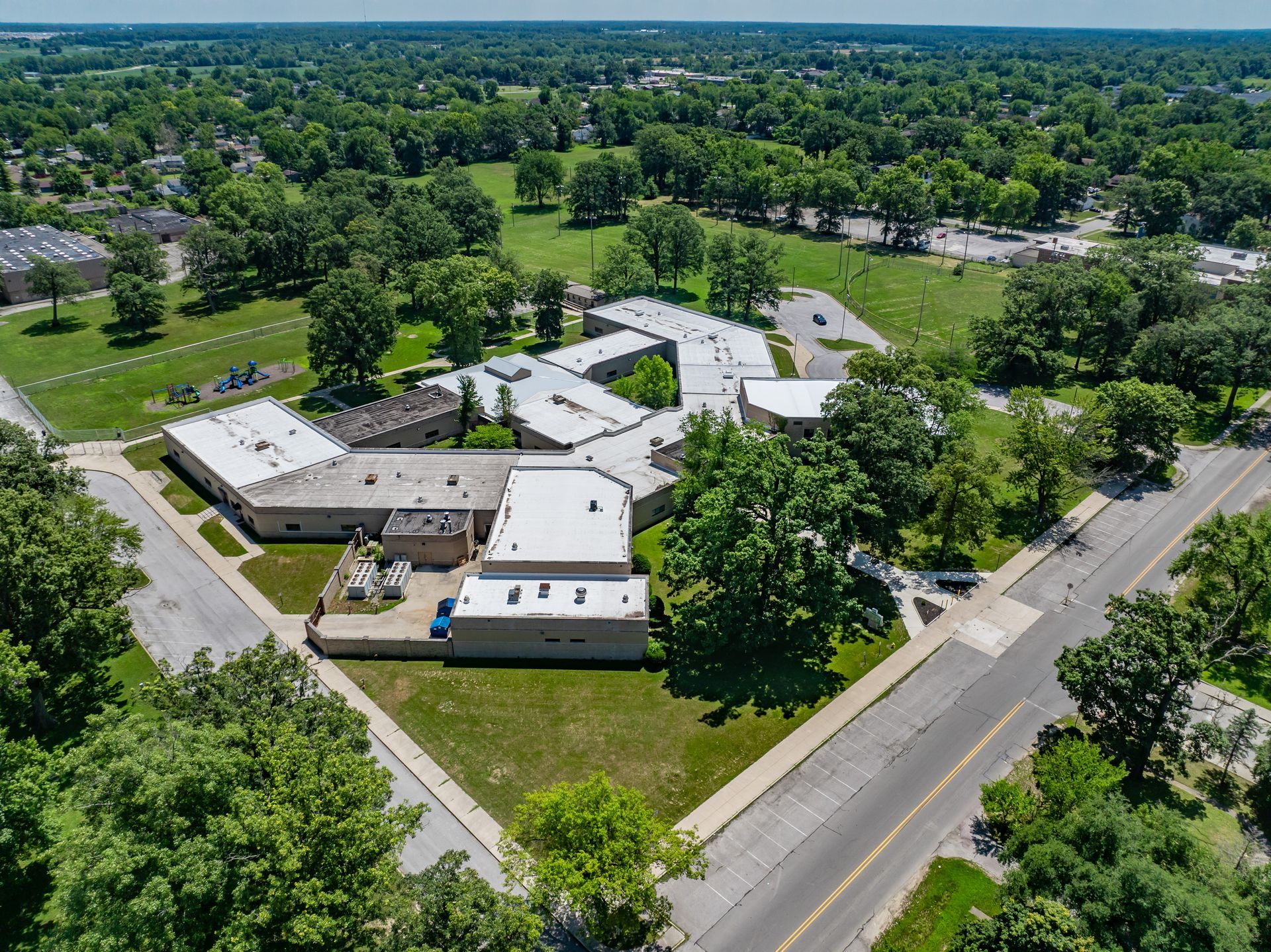 Aerial view of a large white building complex surrounded by trees and roads in a green suburban area