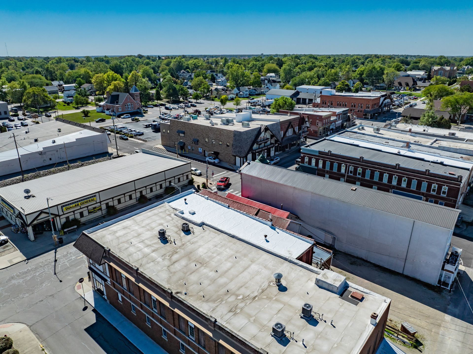 Aerial view of a commercial district with flat-roofed buildings, parking lots, and trees under a clear blue sky