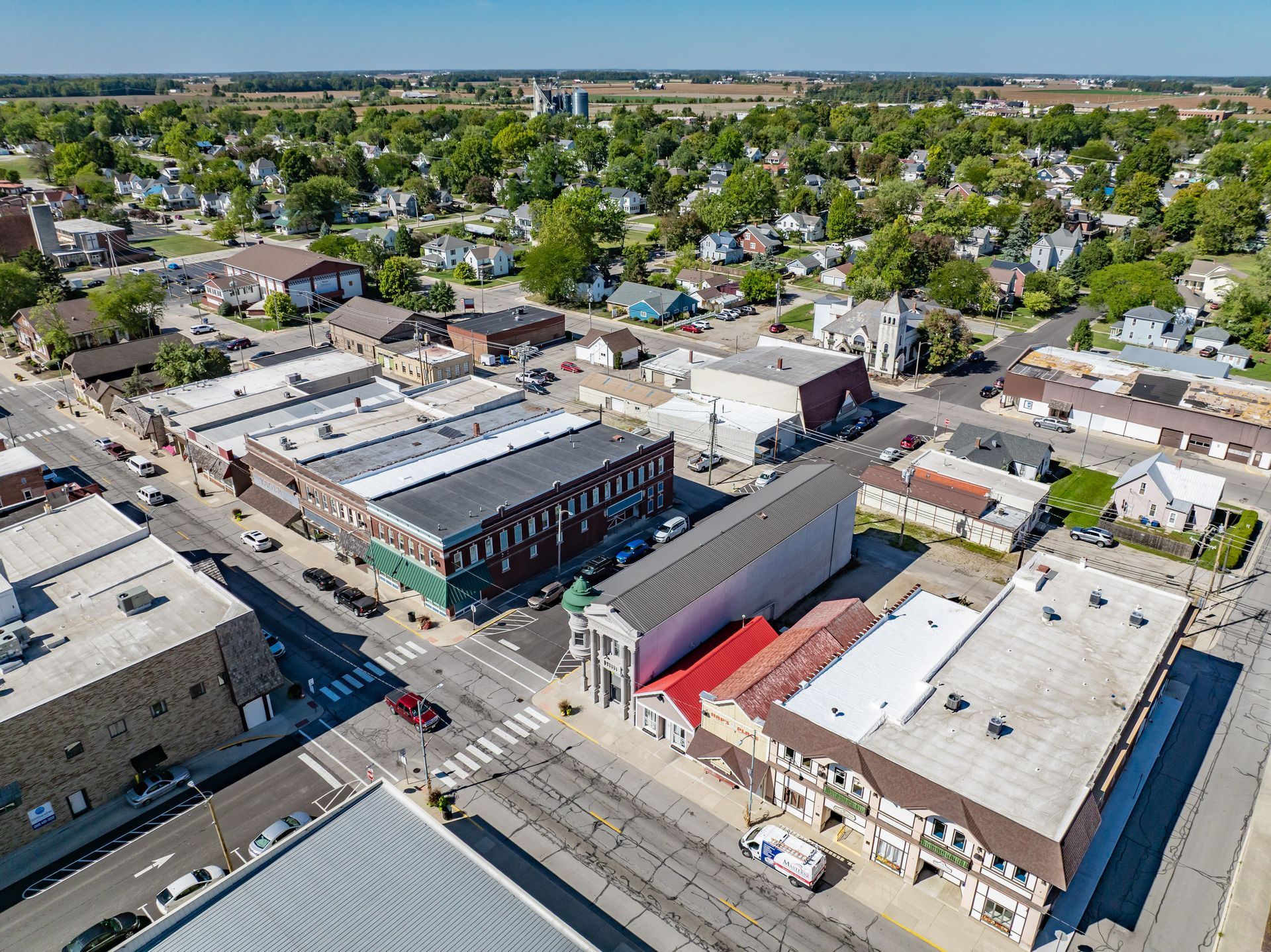 Aerial view of a small downtown block with brick buildings, streets, parked cars, and nearby homes under clear skies