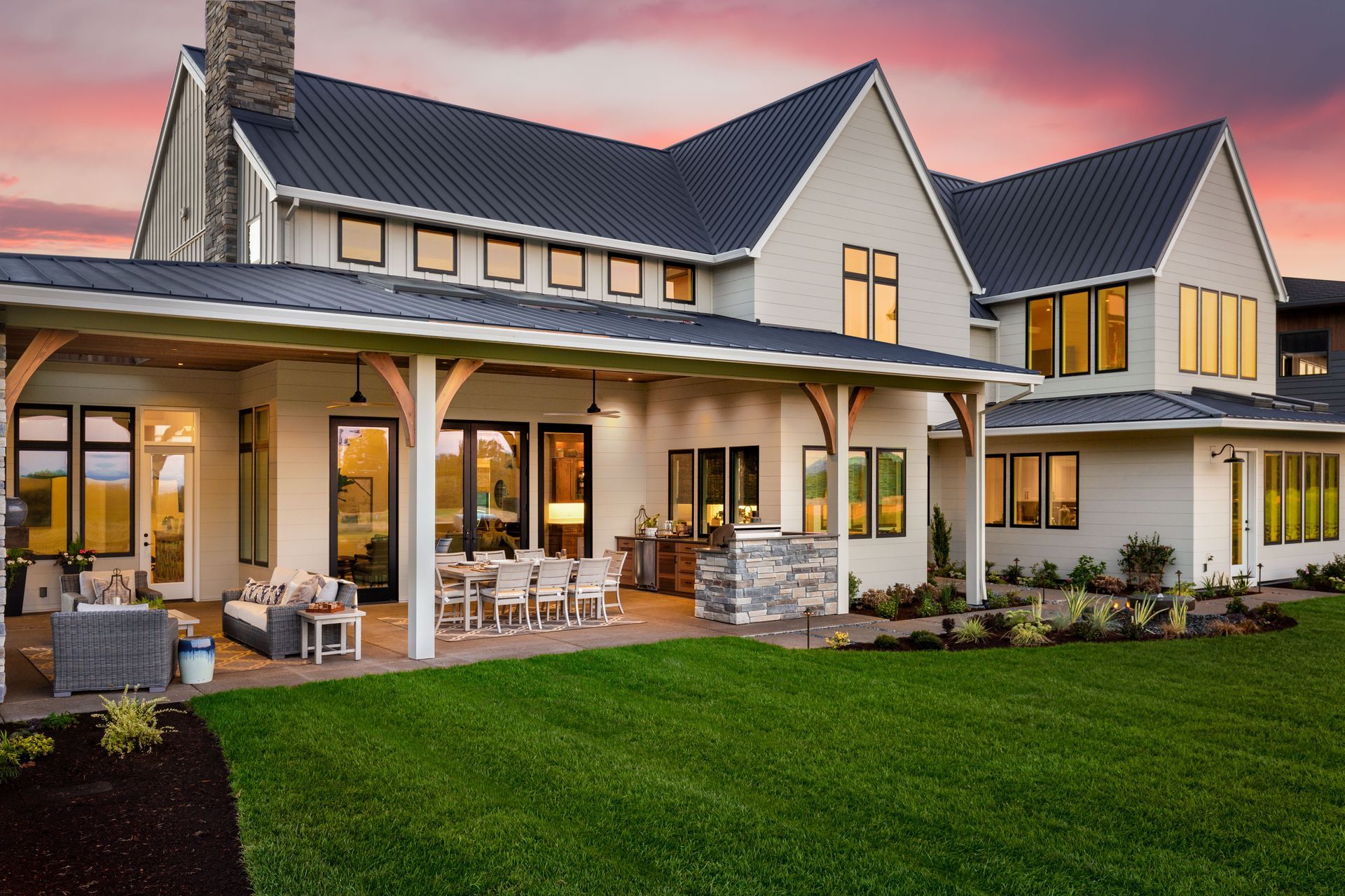 Modern two-story farmhouse with a lit wraparound porch and manicured green lawn at sunset
