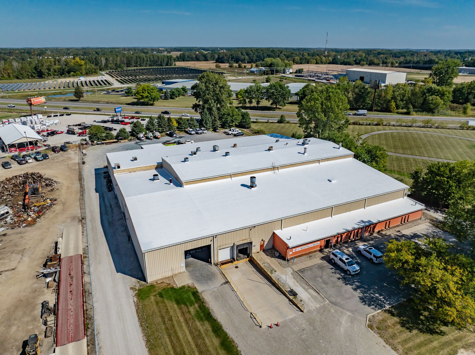 Aerial view of a large white-roofed warehouse beside parking, trees, and open fields
