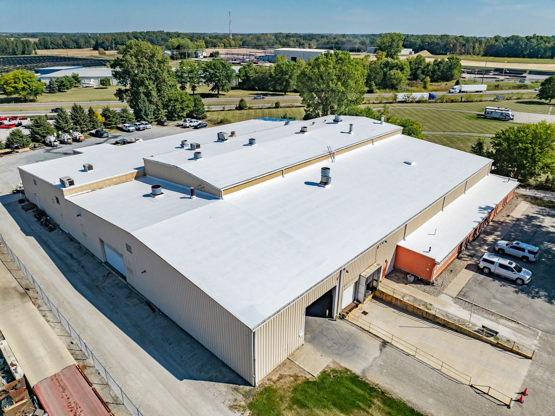 Aerial view of a large white industrial warehouse with rooftop vents, surrounded by parking and trees.