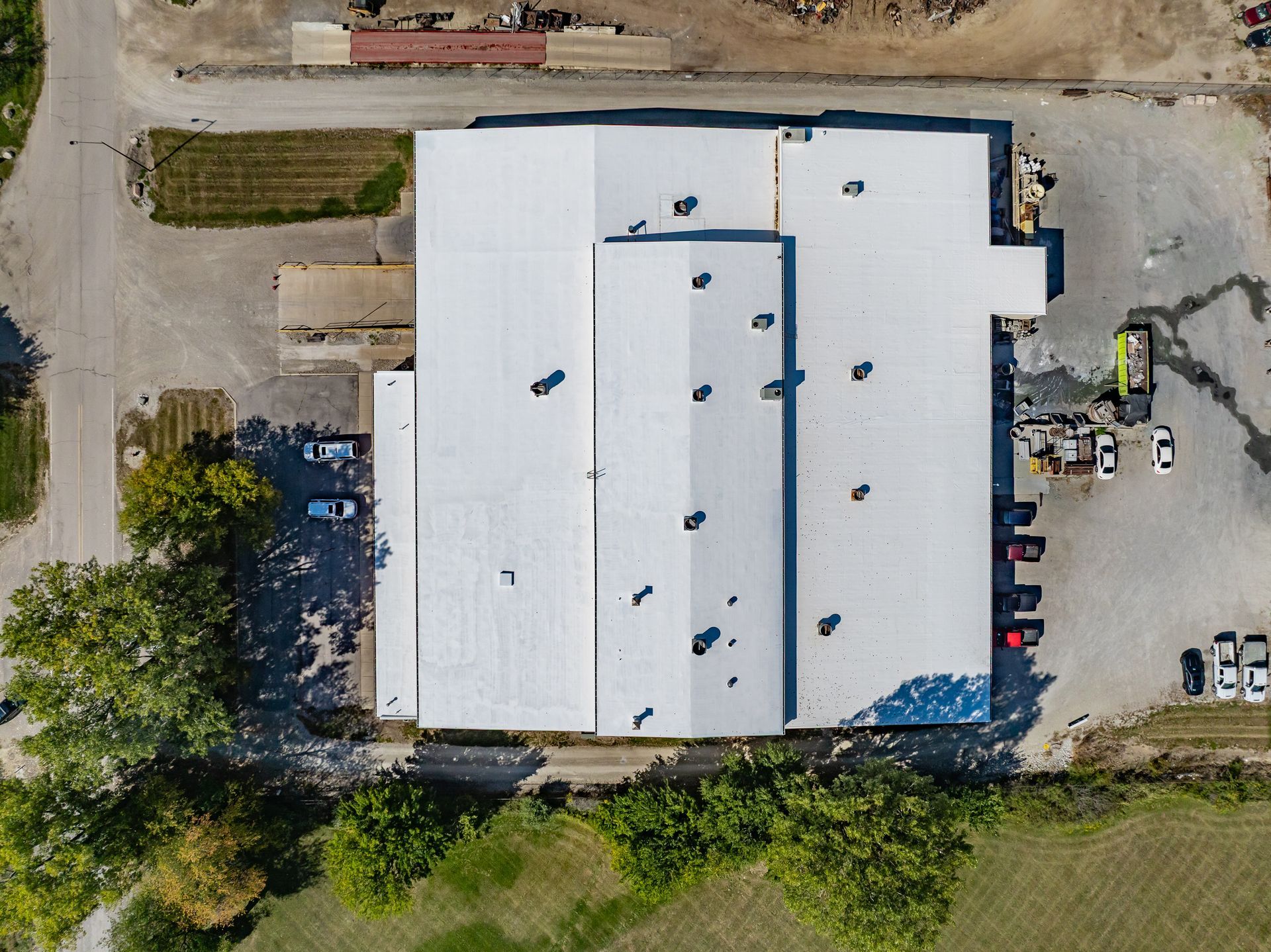 Aerial view of a large white-roofed building with parked cars and surrounding trees