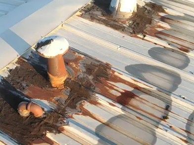 Rusted rooftop vent and pipes on a corrugated metal roof with rust stains and shadows