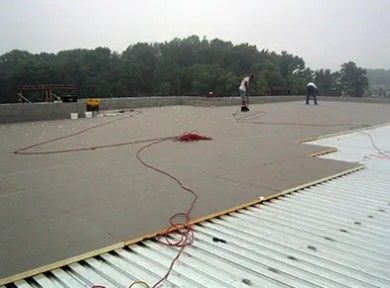 Flat rooftop with workers, cables, and a red rope coil near the edge, trees in the background.