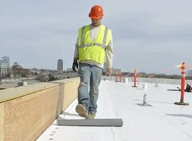Construction worker rolling roofing membrane on a flat rooftop, wearing a neon vest and orange hard hat.