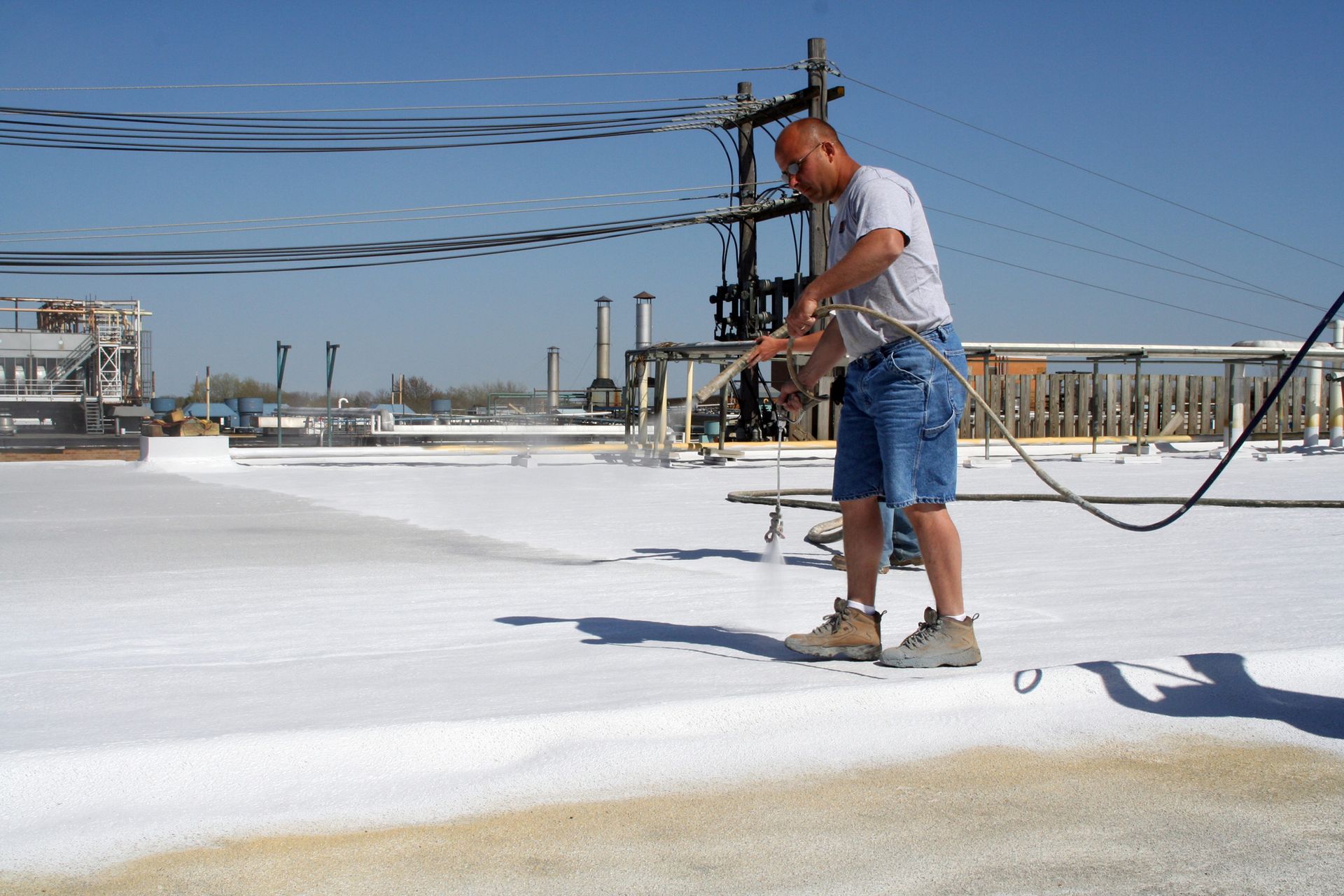 Worker spraying white coating on a flat rooftop with industrial structures in the background