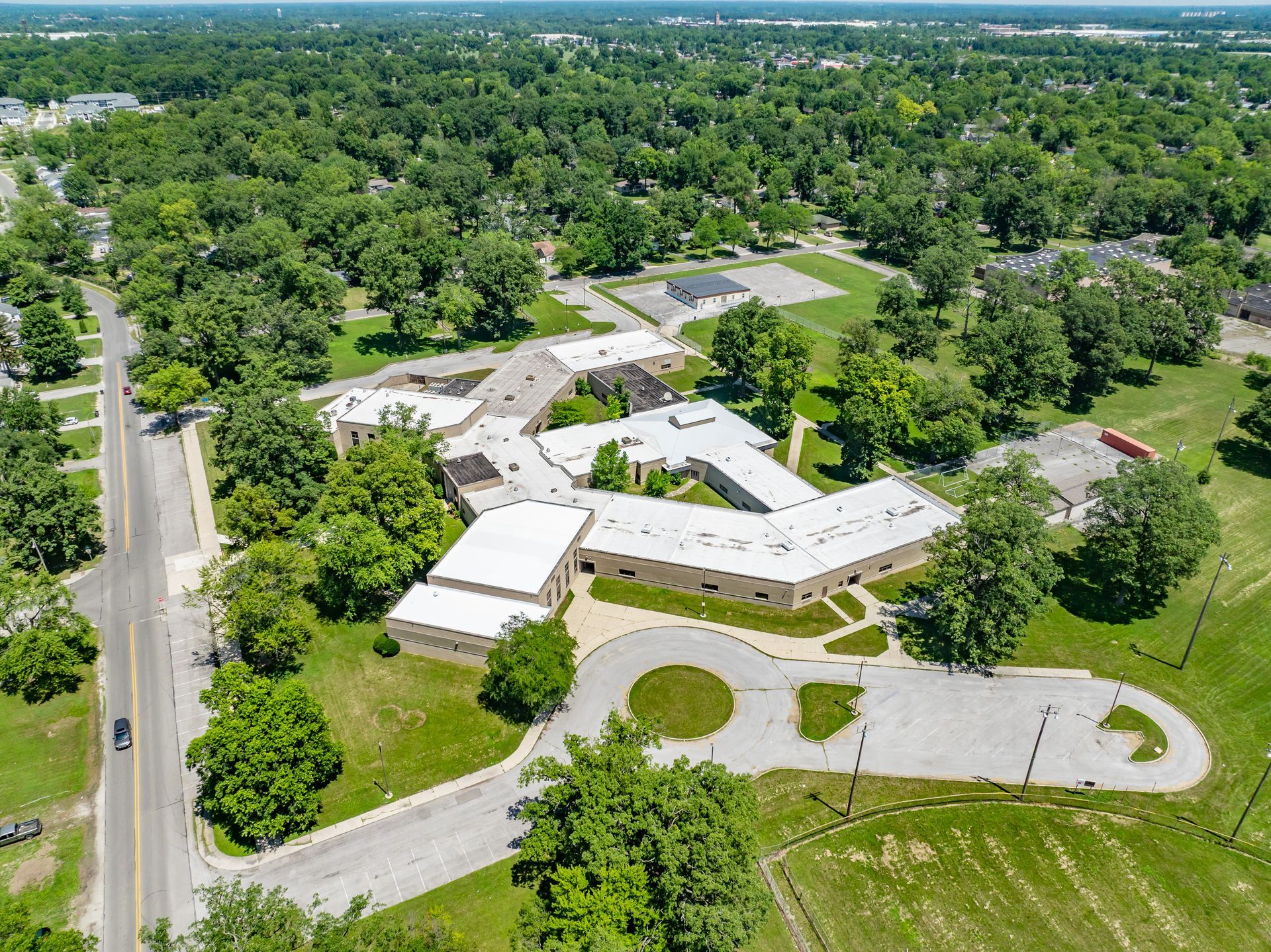 Aerial view of a white-roofed building complex surrounded by trees and roads in a green suburban area