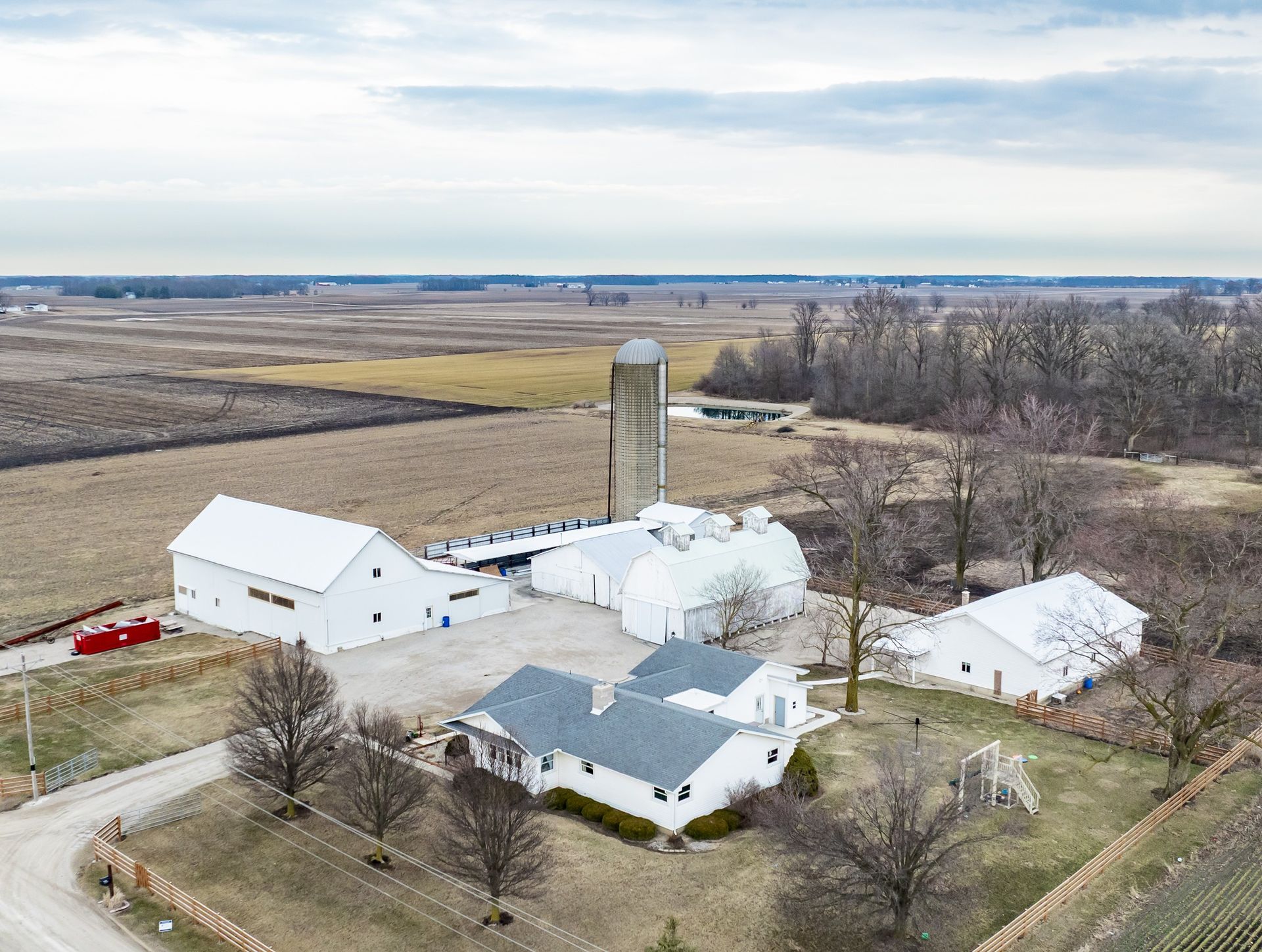 Aerial view of a rural farmstead with white barns, house, silo, and open fields under a clear sky.