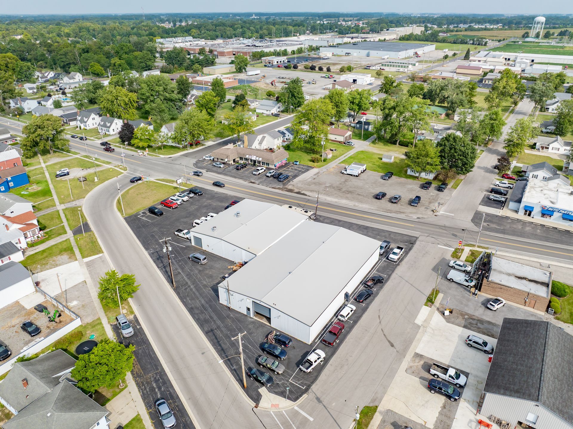 Aerial view of a commercial strip with a large white-roofed building, parking lots, and surrounding trees.