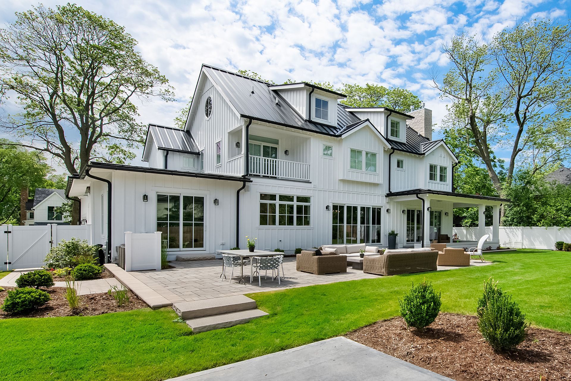 White two-story house with patio seating, green lawn, and trees in a sunny yard
