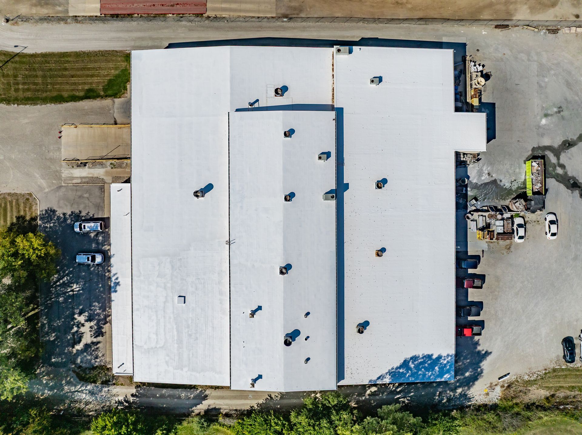 Aerial view of a large white-roofed industrial building with parked cars and equipment around it.