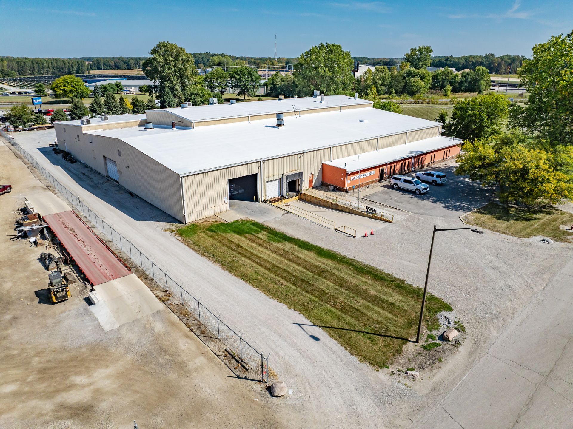 Aerial view of a large beige warehouse with orange loading docks, gravel lot, and surrounding trees