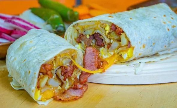 A breakfast burrito cut in half, revealing eggs, cheese, potatoes, and bacon, served with sliced radishes and peppers.