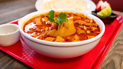 A bowl of vibrant red menudo soup topped with cilantro, served on a red tray with a side of tortillas and lime.