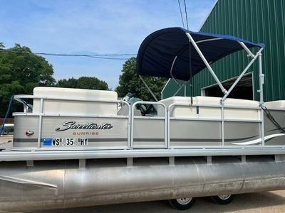 Pontoon boat with blue canopy docked on a trailer outdoors, with silver fencing and green trees behind it