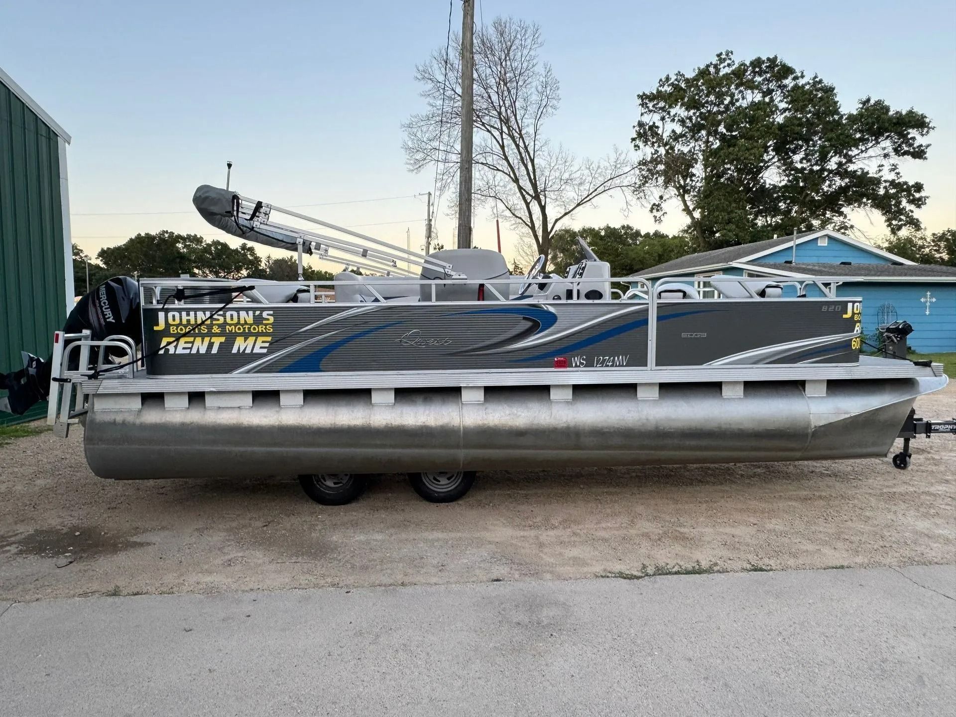 Pontoon boat on a trailer in a gravel lot, with a raised motor and trees in the background