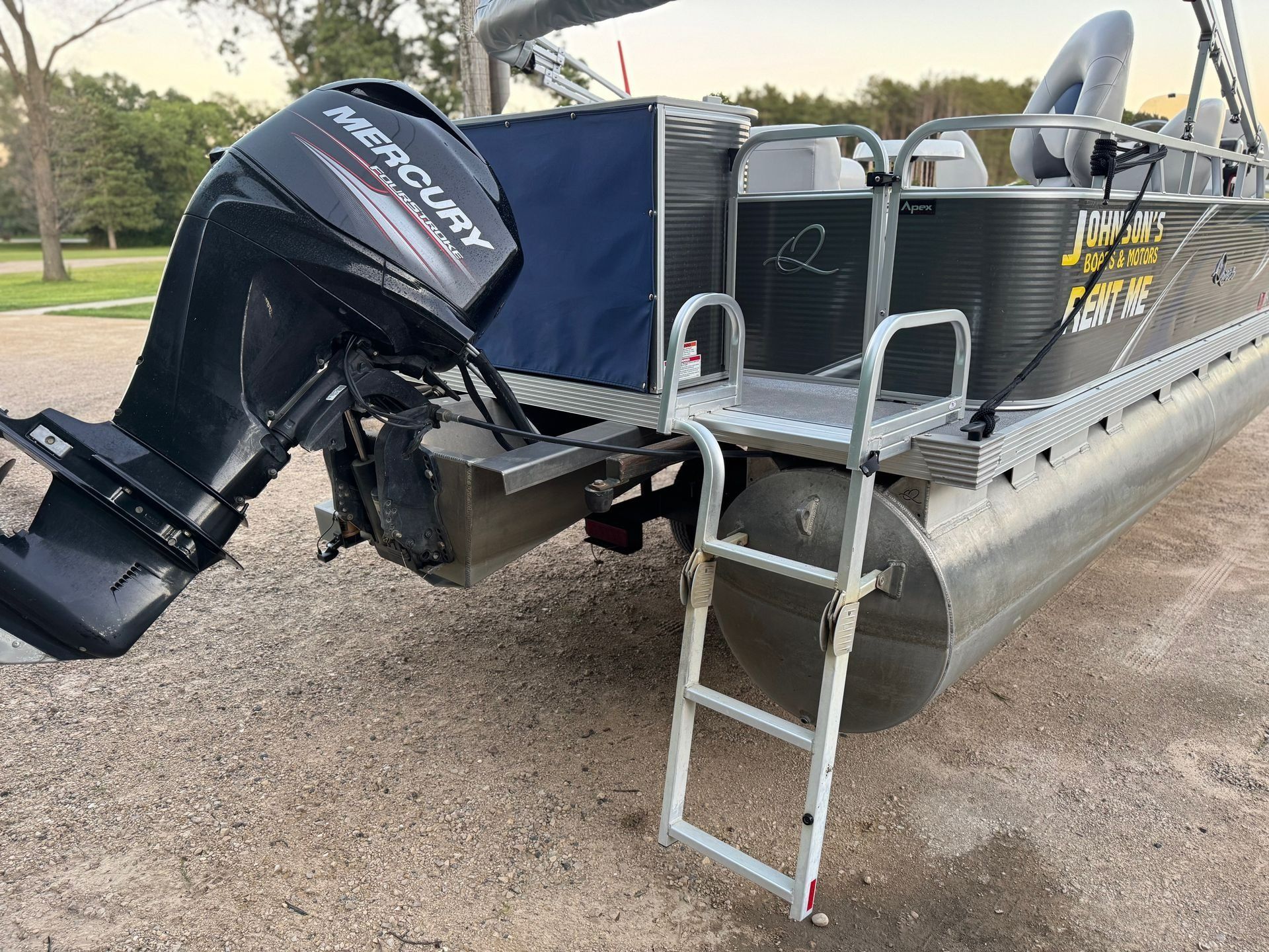 Pontoon boat with black Mercury outboard motor docked on gravel, with a metal boarding ladder attached.