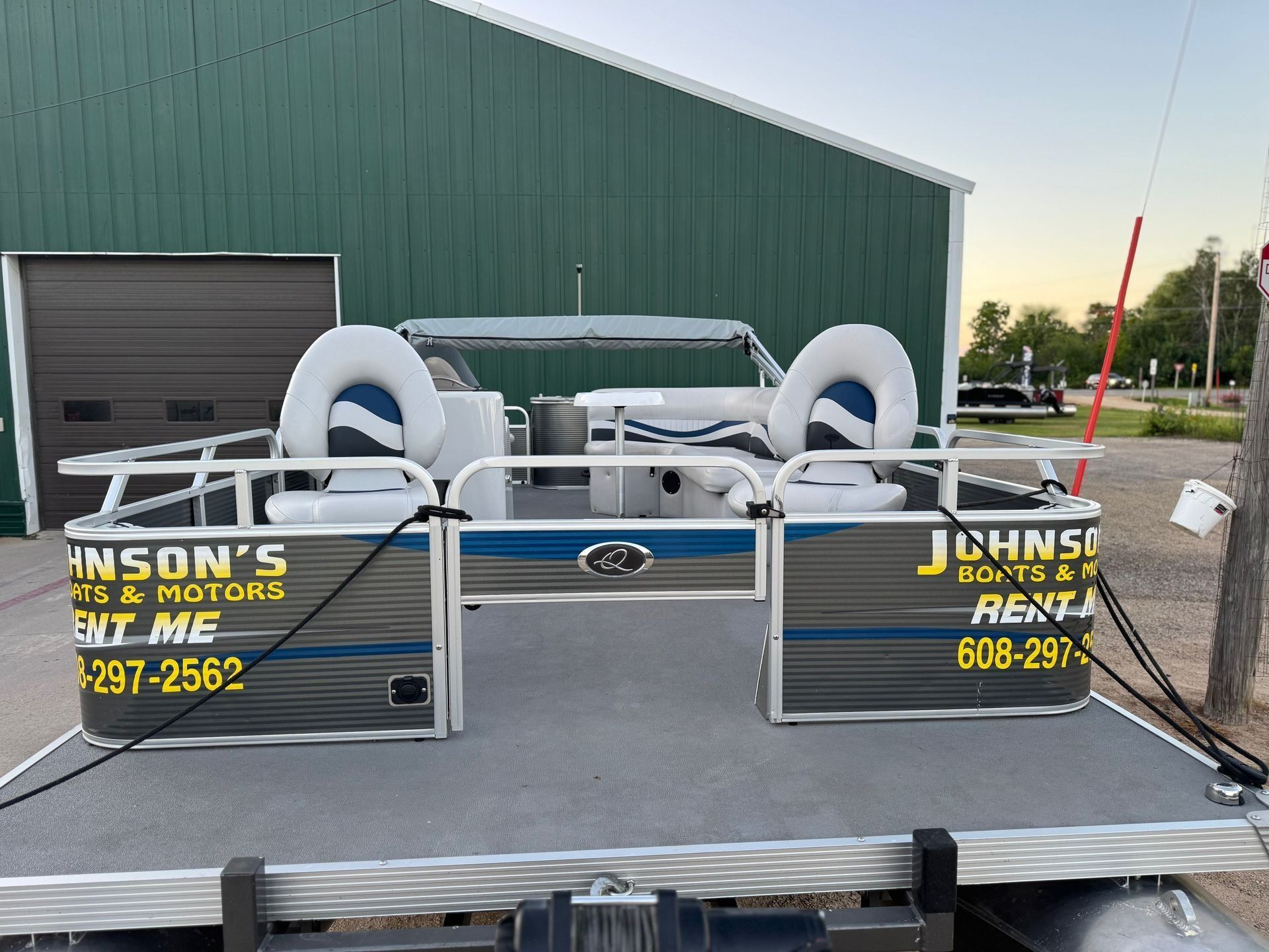 Pontoon boat on a trailer with white seats, parked beside a green building with signs and coiled ropes.