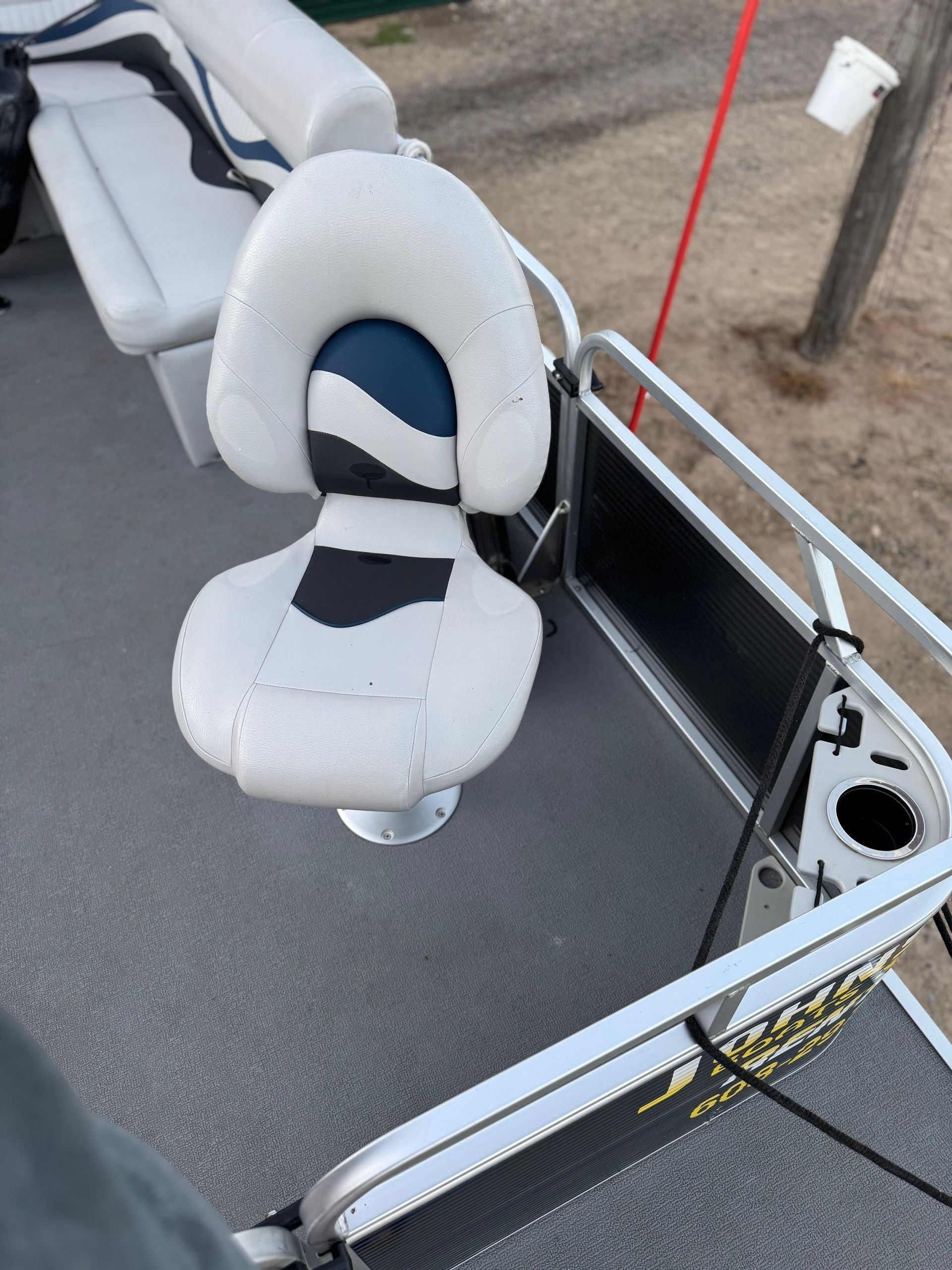 White padded boat seat with blue center mounted on a deck beside a metal railing and sandy shore