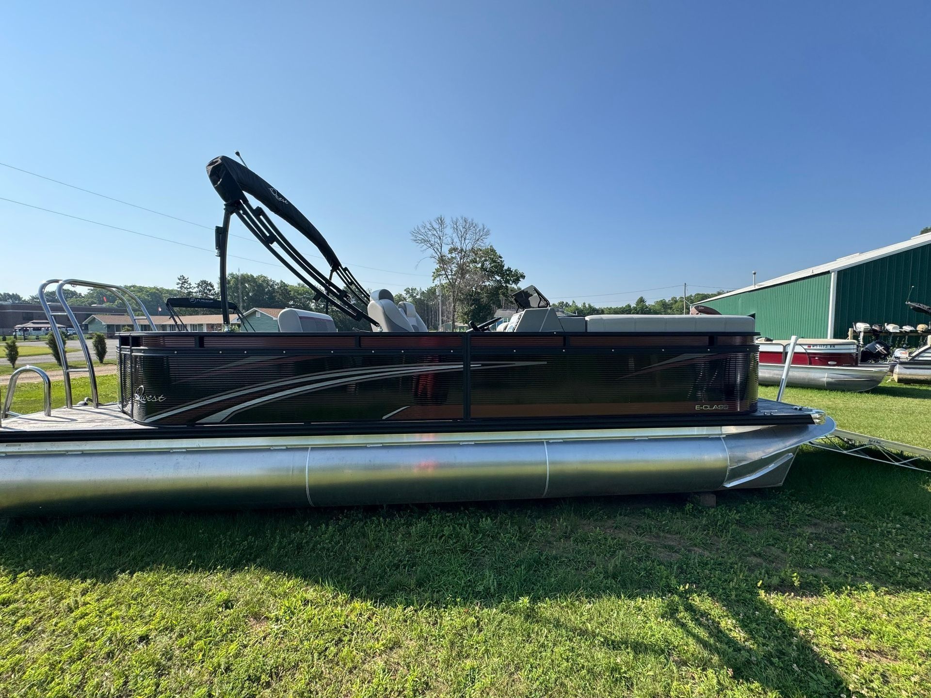 Pontoon boat with a black crane attachment on a grassy lot under a clear blue sky