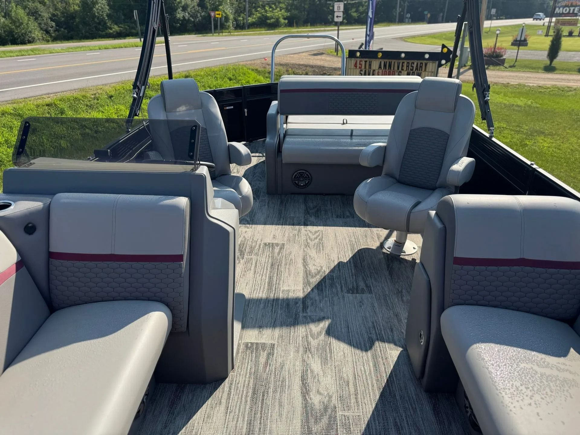 Pontoon boat interior with gray cushioned seats on a lake trailer, parked by a road and trees.