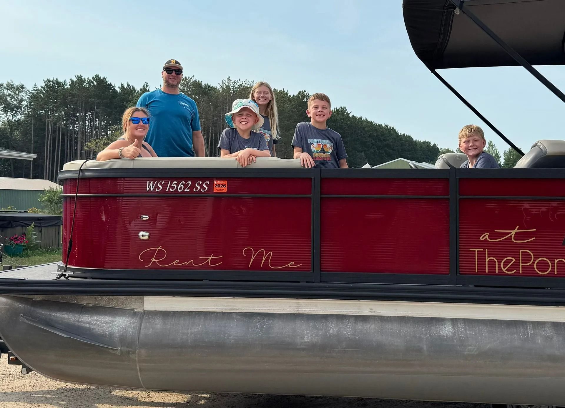 Four people ride in a red pontoon boat on a lake, smiling and waving