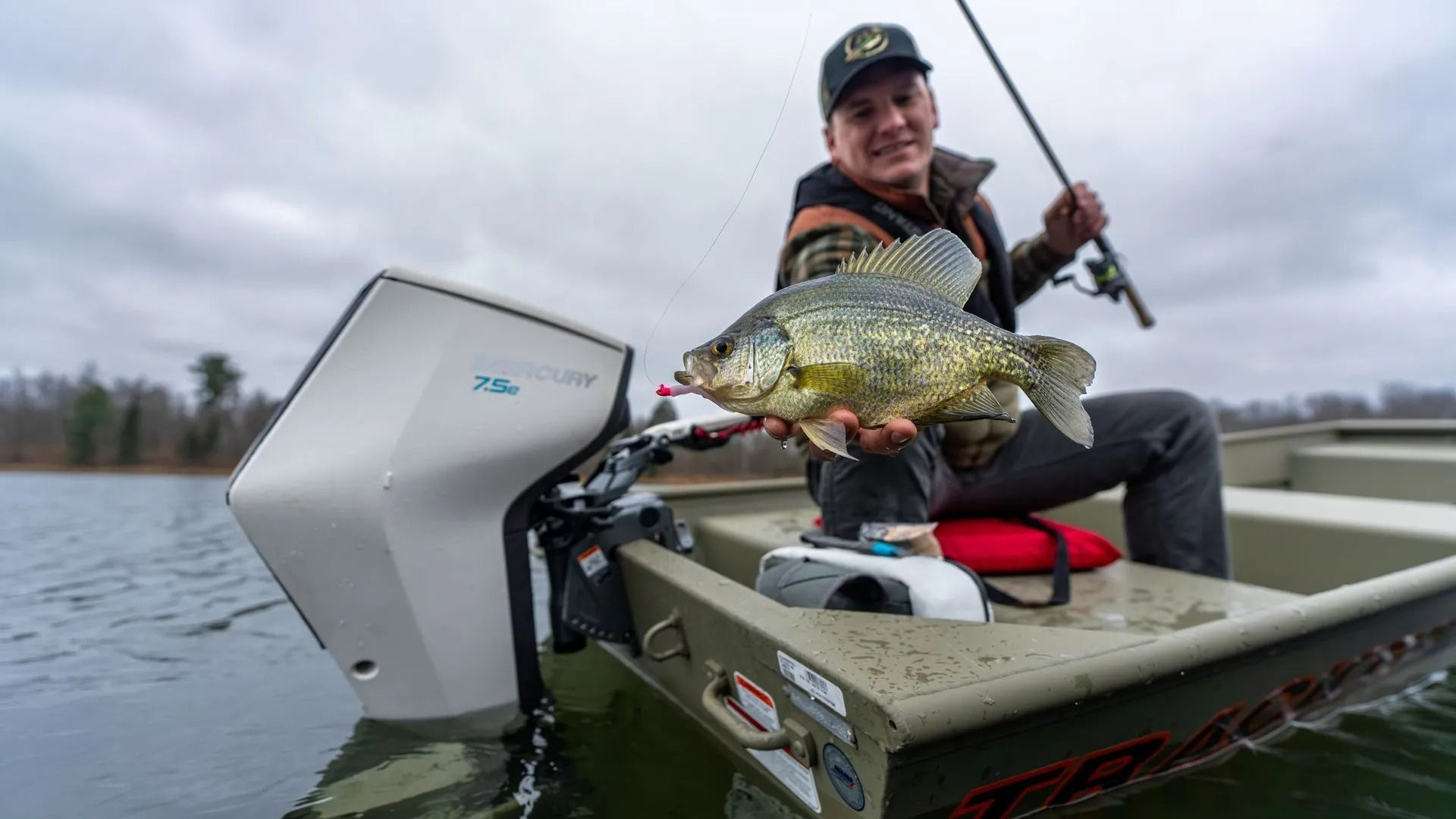 Angler on a boat holds a large fish over a calm lake under an overcast sky